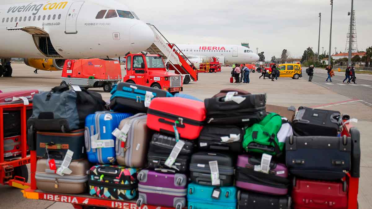 Equipaje facturado acumulado en la pista de un aeropuerto junto a aviones de aerolíneas comerciales