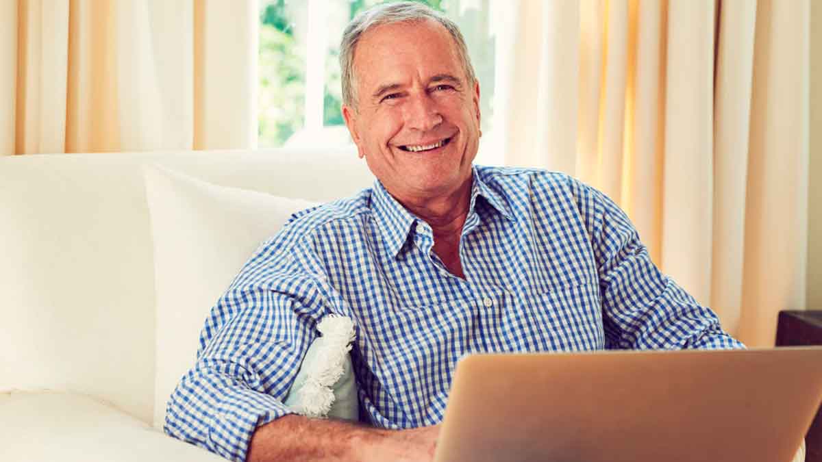 Jubilado sonriente usando un ordenador portátil en casa tras la subida de la pensión en 2026