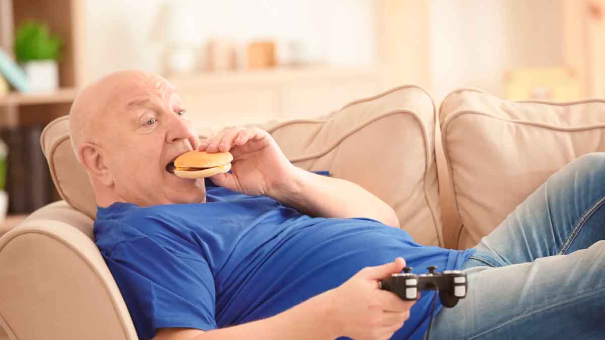 Hombre adulto sentado en el sofá comiendo una hamburguesa, ejemplo de hábitos sedentarios que afectan a la salud