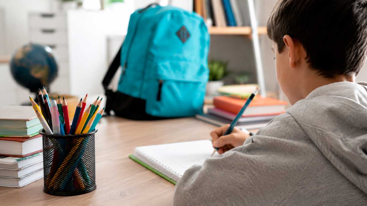 Alumno escribiendo en un cuaderno con libros, lápices y mochila escolar sobre una mesa de estudio