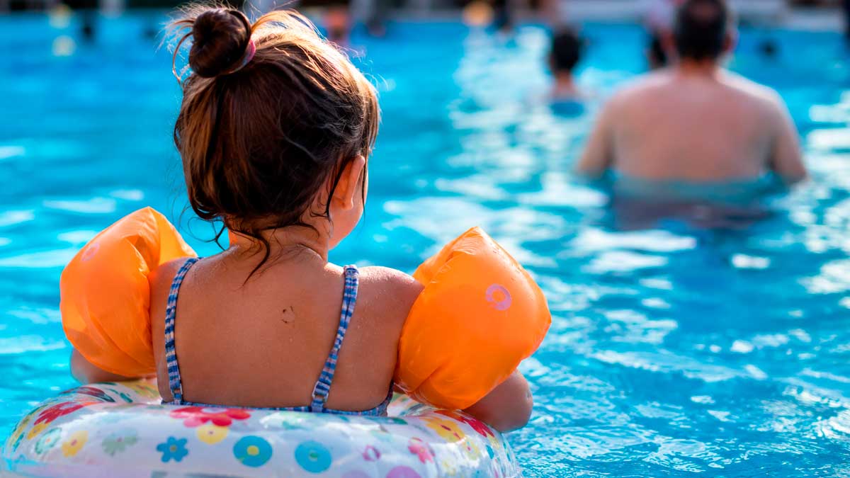 Niña con manguitos y flotador en una piscina bajo la supervisión de un adulto