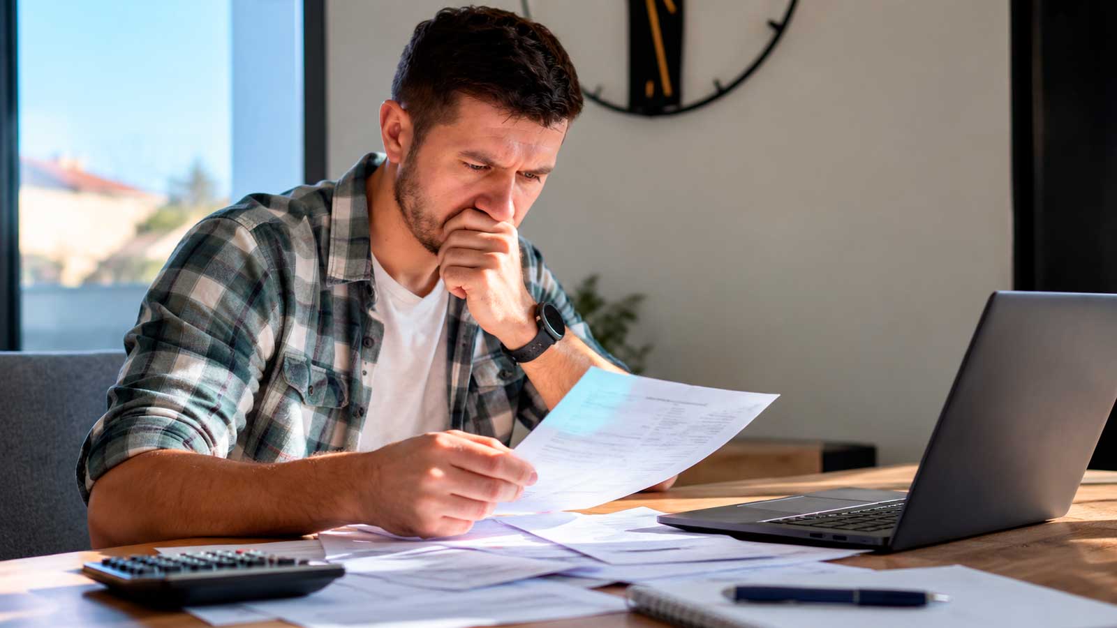 Trabajador preocupado revisando nóminas impagadas y documentos salariales frente a su ordenador portátil.